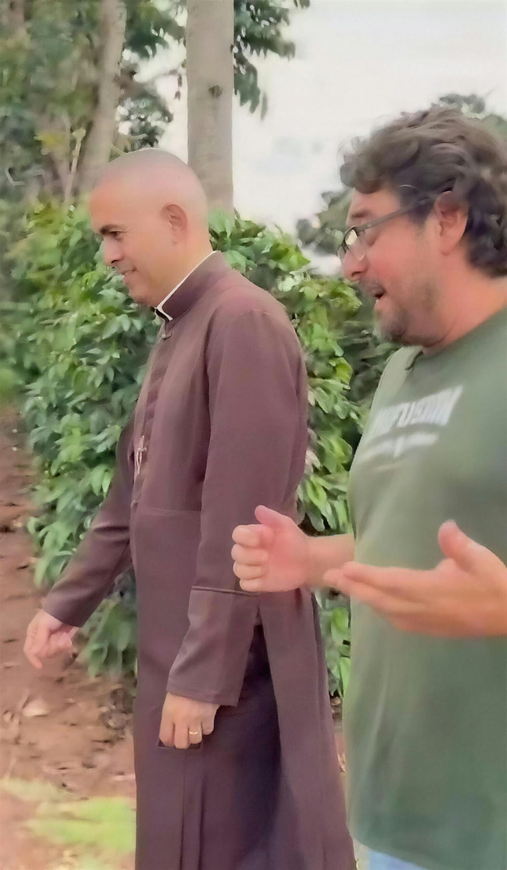 Um padre de batina marrom e o produtor conversam enquanto caminham entre as fileiras de café